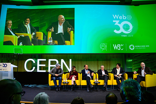 Six panelists sitting in yellow chairs engaging one another on stage at the Web 30 celebration.