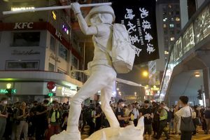 A nighttime photo of a white statue holding a folded umbrella and flag that reads "Free Hong Kong" on a crowded city street.