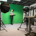 Person standing in front of green screen and behind cameras and lighting on tripods in a studio space