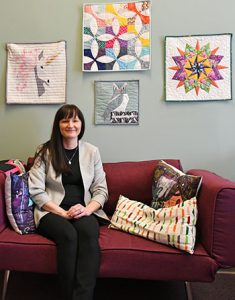 Casey sits on a burgundy couch in her office, surrounded by colorful pillows and quilted wall hangings.