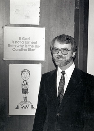 Photo of Fred Roper next to a door with a sign saying "If God is not a Tarheel, then why is the sky Carolina Blue!" and a cartoon.