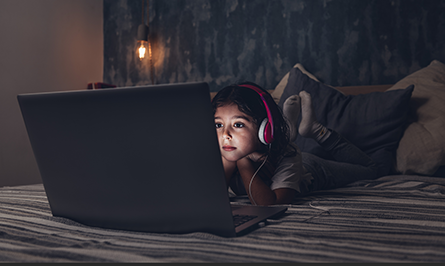 Young girl wearing headphones and lying on a bed looking at the screen of a laptop